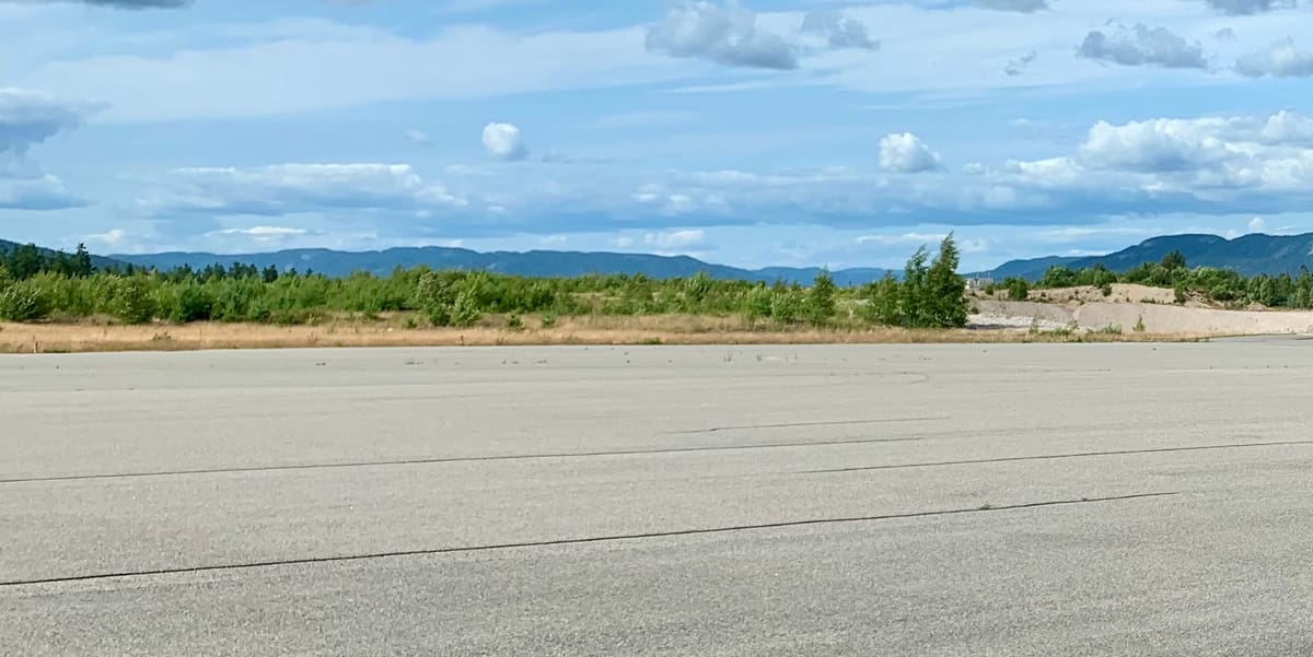 Bred asfaltflate under sommerhimmel, med lav skog og blå åser i horisonten